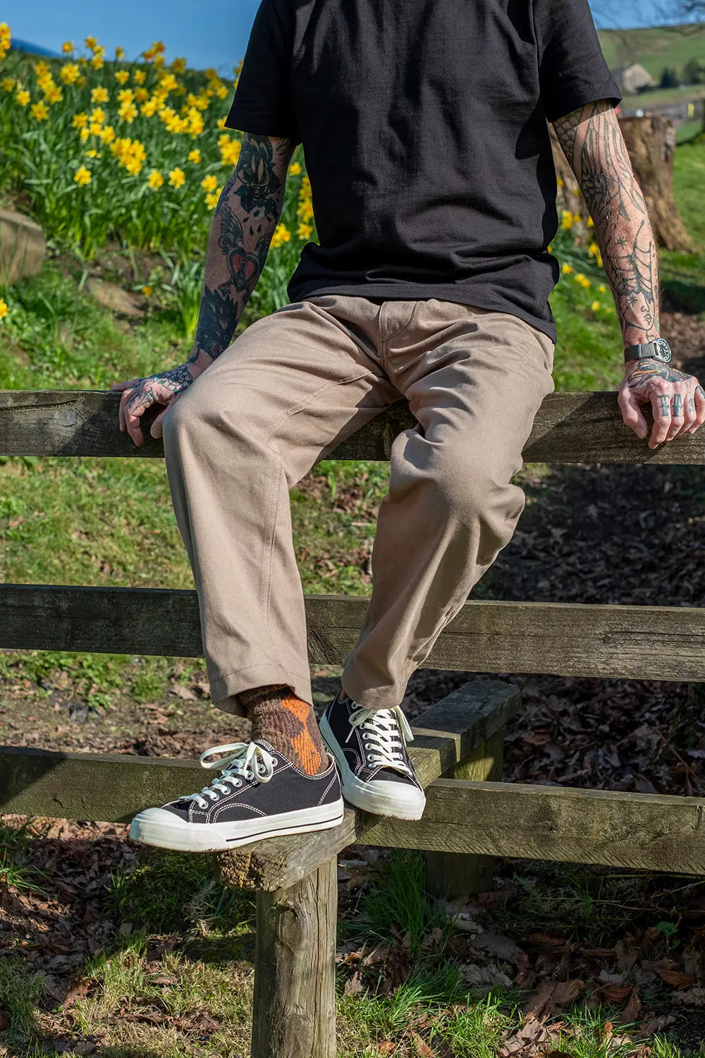 A person sitting on a wooden fence outdoors, wearing beige straight-leg trousers, a black t-shirt, and black canvas sneakers with white soles. The background features a grassy field with yellow wildflowers under a blue sky.
