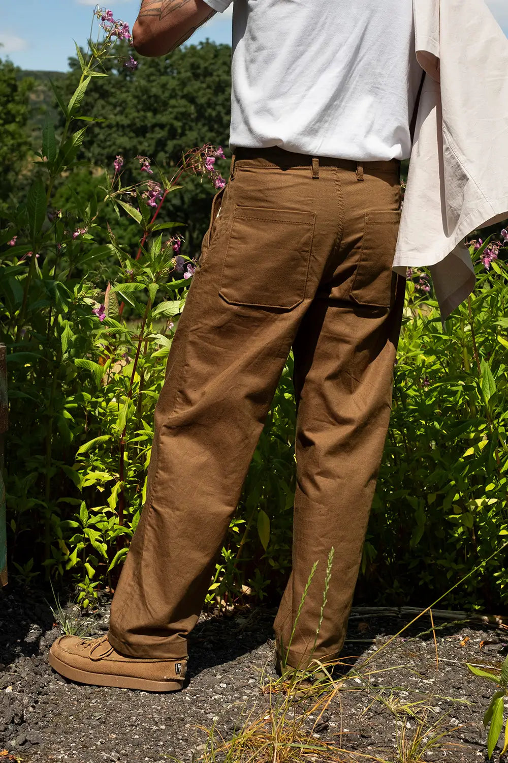 A person standing outdoors in a garden wearing brown work pants, a white t-shirt, and tan work boots, with green foliage in the background.