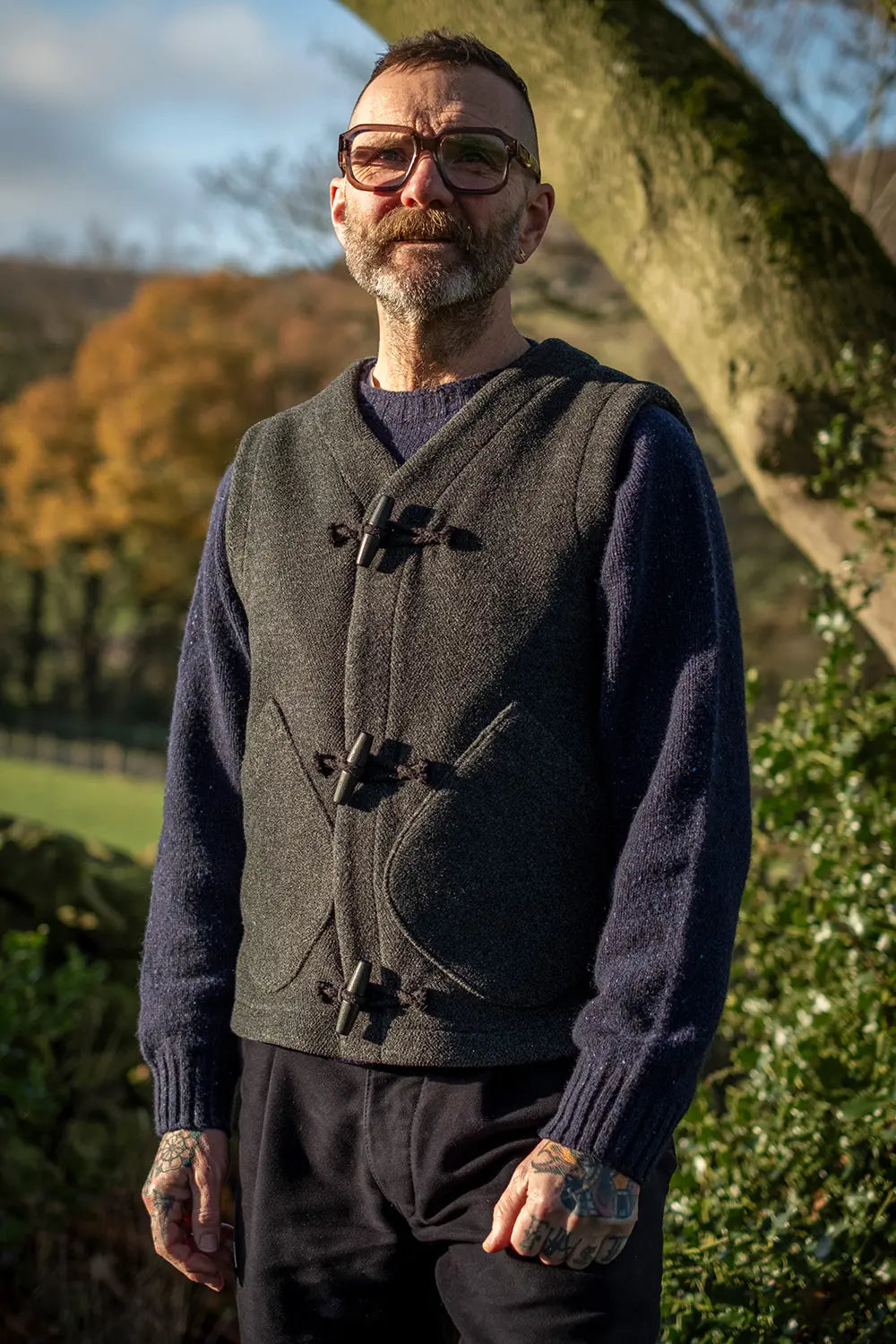 A man wearing a dark gray wool teasel duffel  vest over a navy blue knit jumper, standing outdoors in front of a tree with autumn foliage in the background.