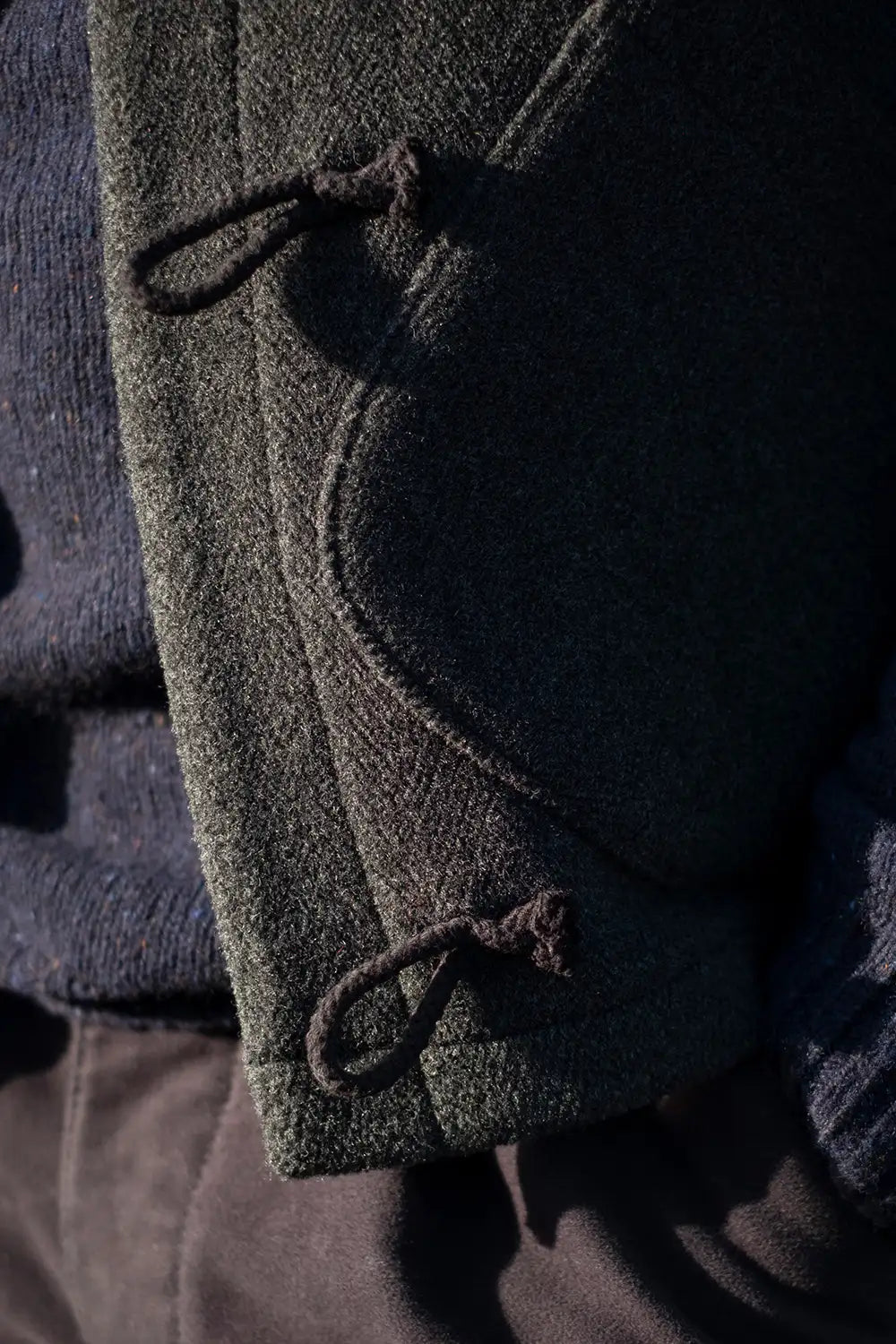 Close-up of a teasel duffel wool vest with dark brown toggle closures, layered over a navy knit sweater.