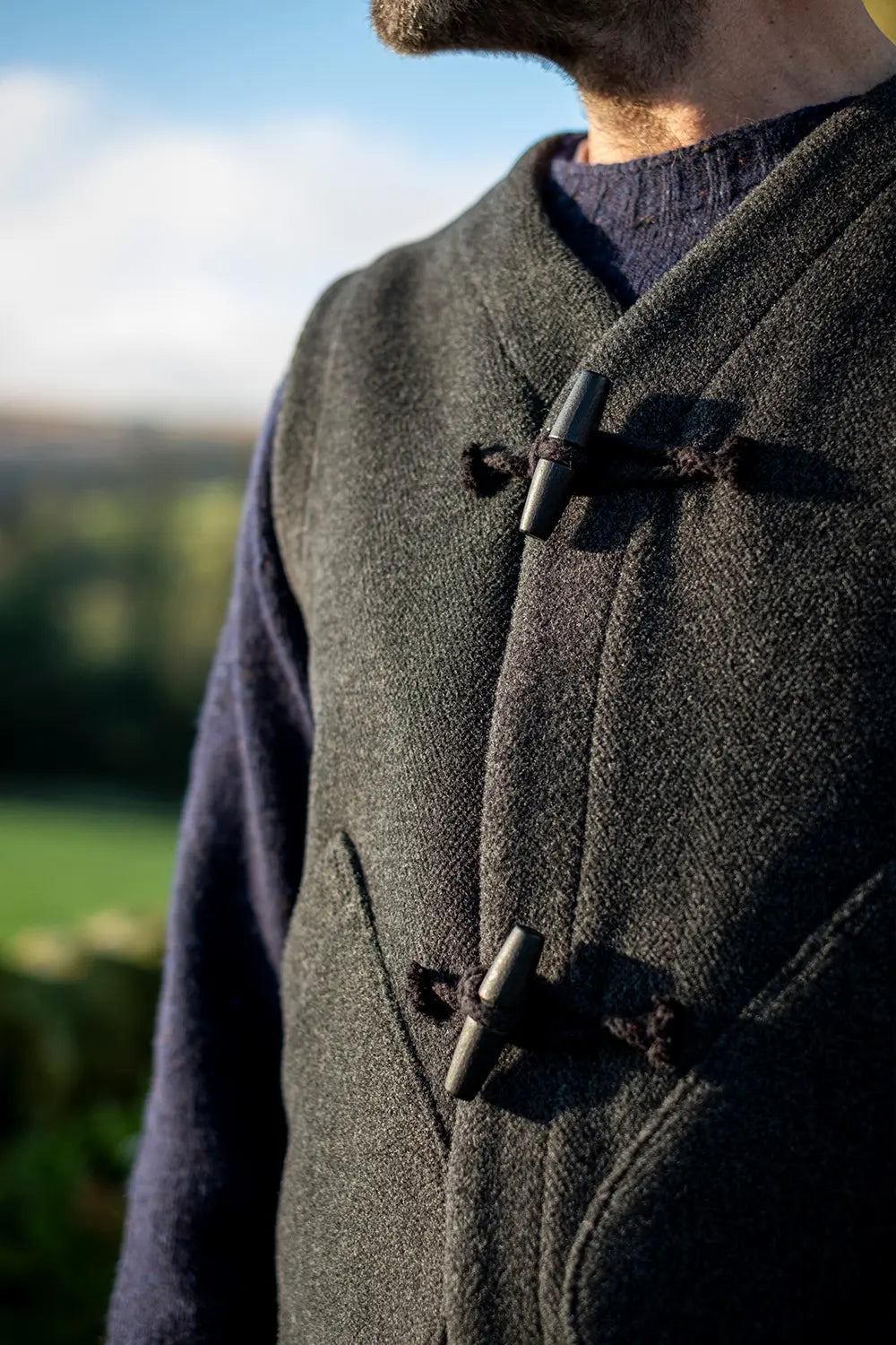 Close-up of a person wearing a dark charcoal wool duffle vest over a navy jumper, with a blurred outdoor landscape in the background.