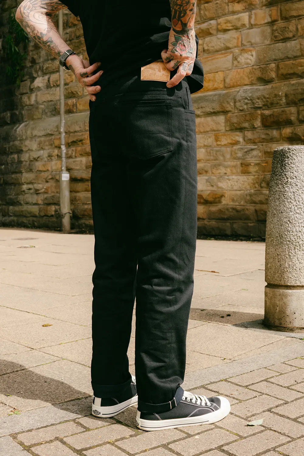 A person wearing black wide-leg trousers, a black t-shirt, and black low-top sneakers with white soles, standing outdoors against a stone wall.
