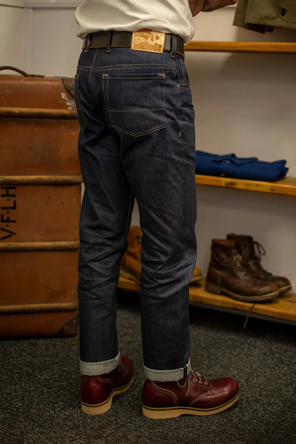 A person shown from the waist down wearing dark blue denim jeans with rolled cuffs, paired with a white shirt, brown belt, and dark red work boots, standing in a workshop setting.