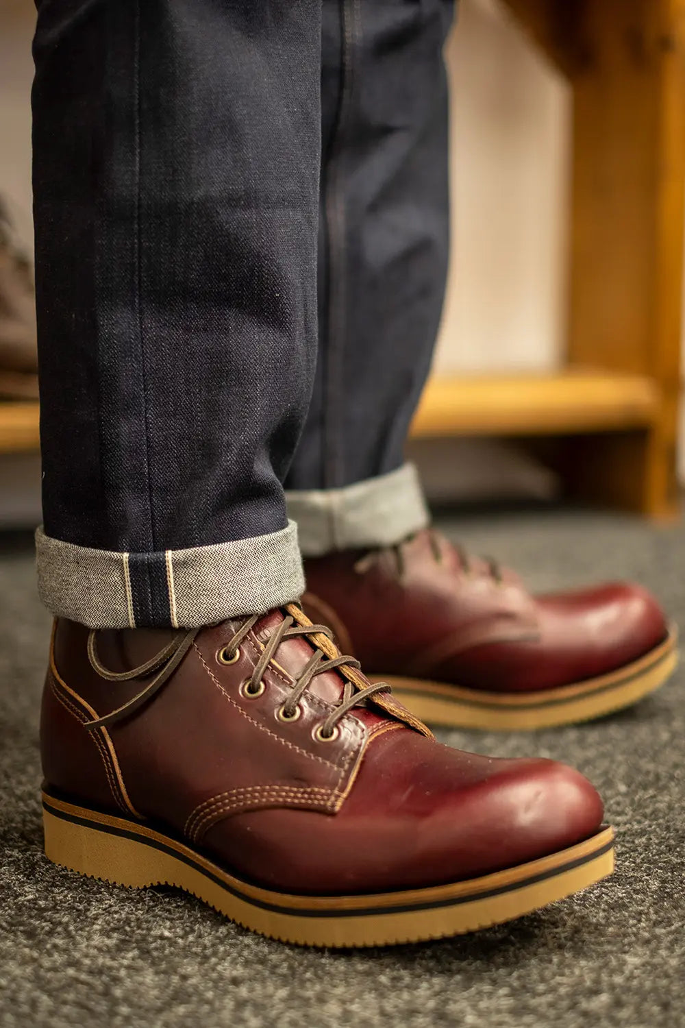 A close-up shot of a person's feet wearing dark brown leather lace-up work boots with tan soles, paired with dark cuffed jeans.