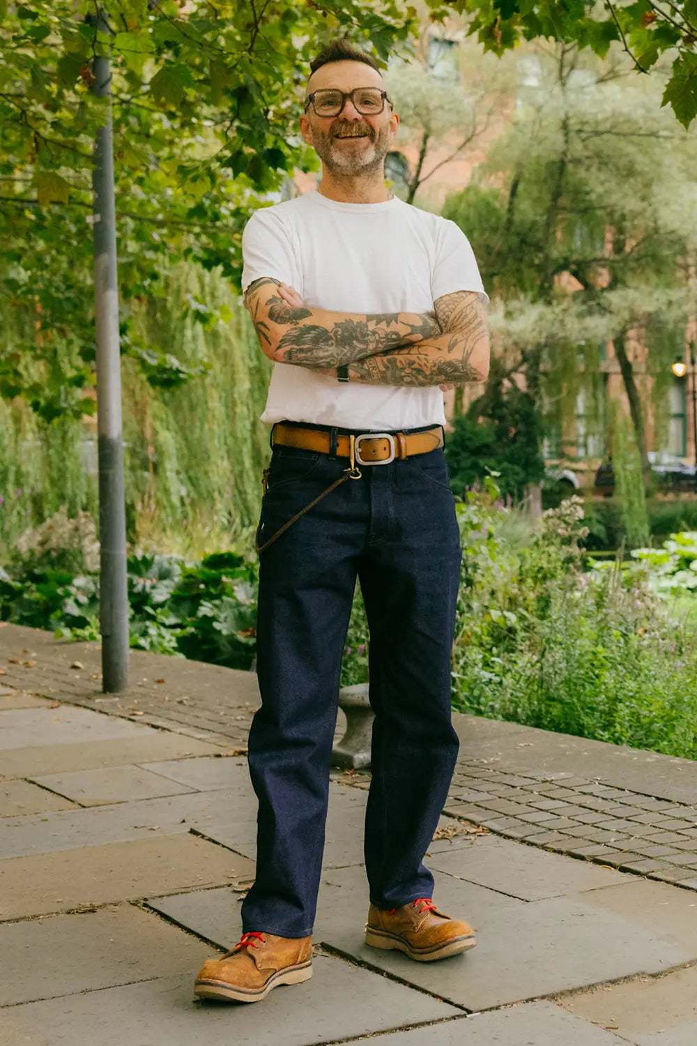 A man with glasses and arm tattoos stands outdoors on a stone walkway wearing a white t-shirt, dark navy blue jeans, a brown leather belt, and tan work boots, with green foliage in the background.