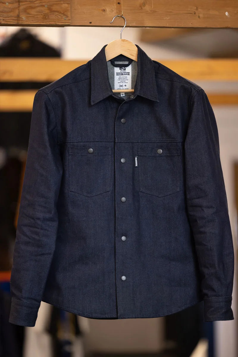 A dark indigo denim long-sleeve button-up shirt hanging on a wooden hanger against a warm, blurred background.