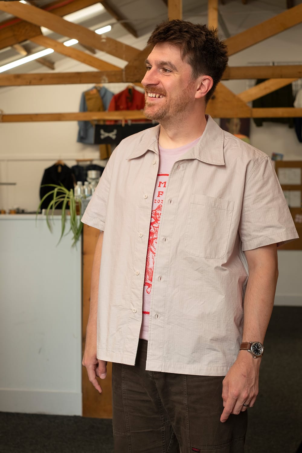 A man wearing a light beige short-sleeve button-up shirt over a pink graphic t-shirt and dark brown pants, standing in a workshop or studio environment with wooden ceiling beams and clothing racks in the background.