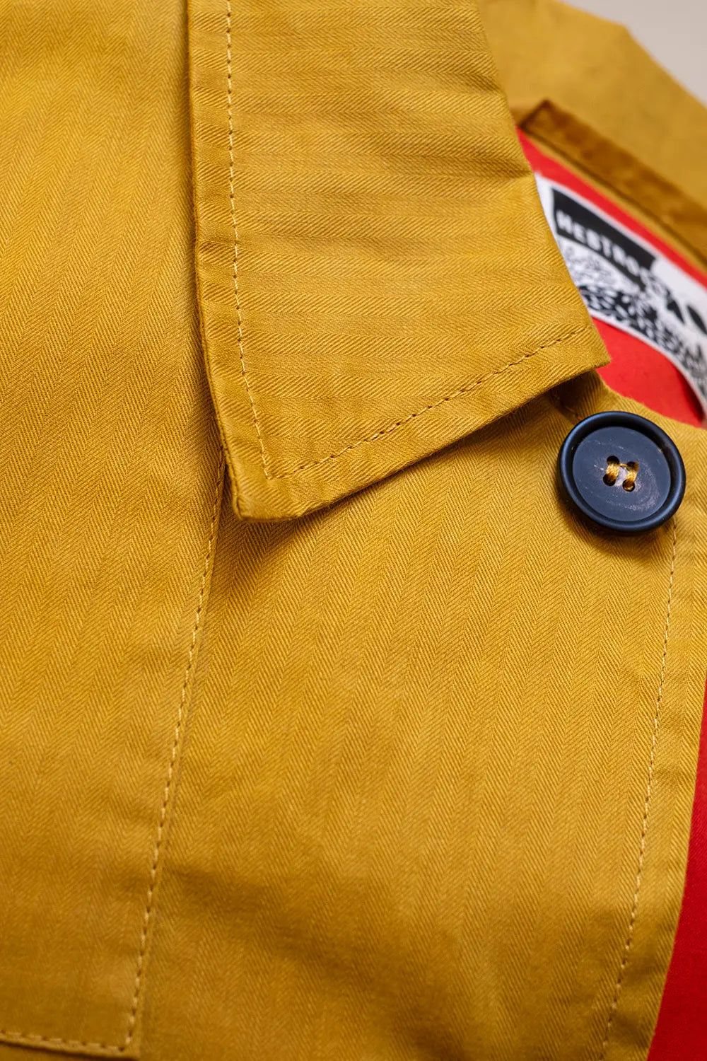 Close-up of a mustard yellow work shirt with a dark navy button and a red inner collar lining.