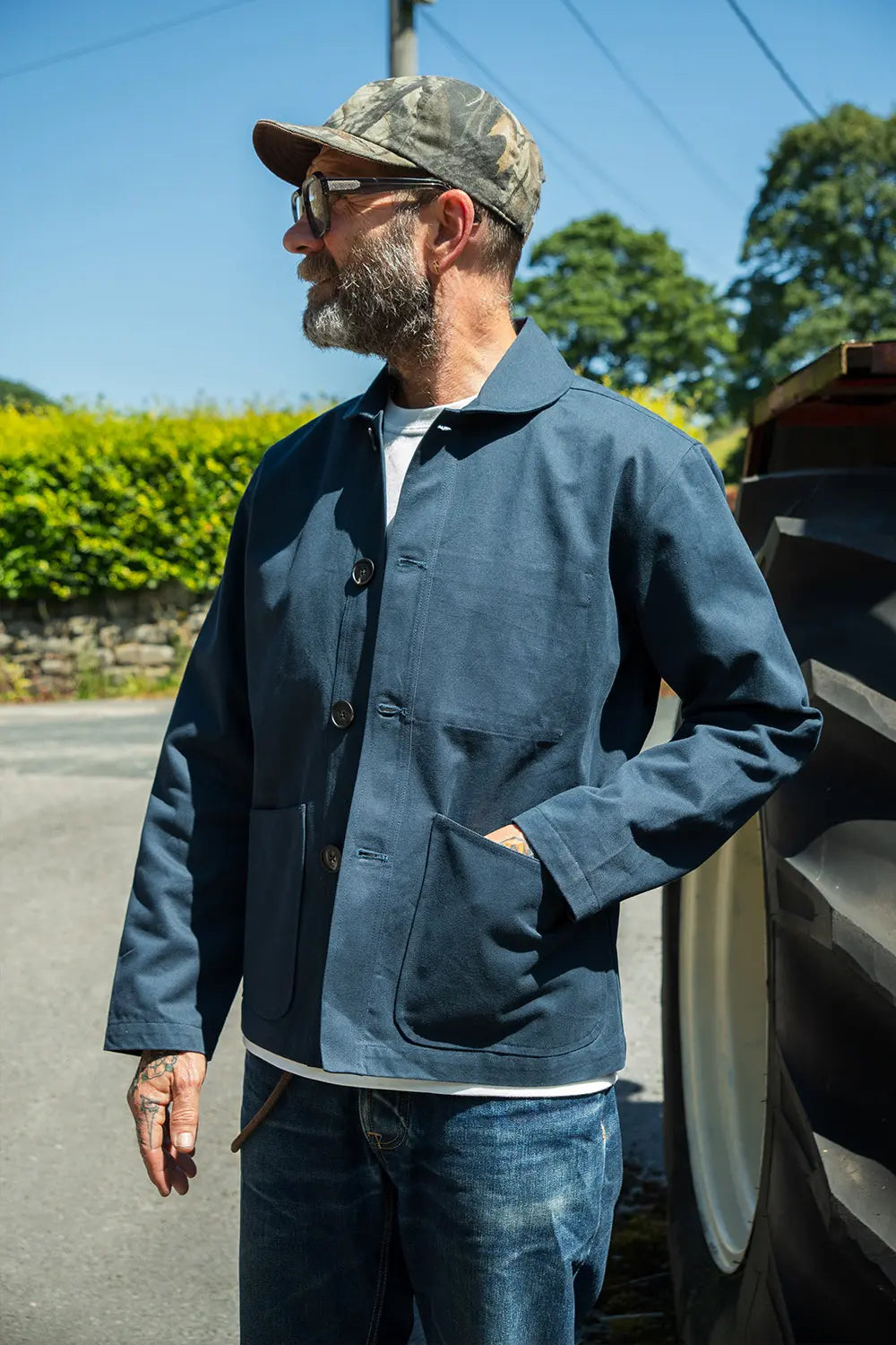 A man wearing a navy blue canvas utility jacket, blue jeans, a camouflage baseball cap, and sunglasses, standing outdoors next to a tractor.