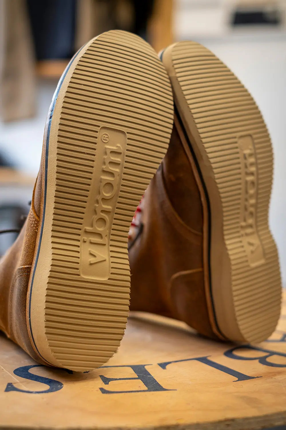 Close-up of the soles of a pair of brown boots, showing the Vibram brand name embossed in the tan rubber outsoles, resting on a wooden surface.