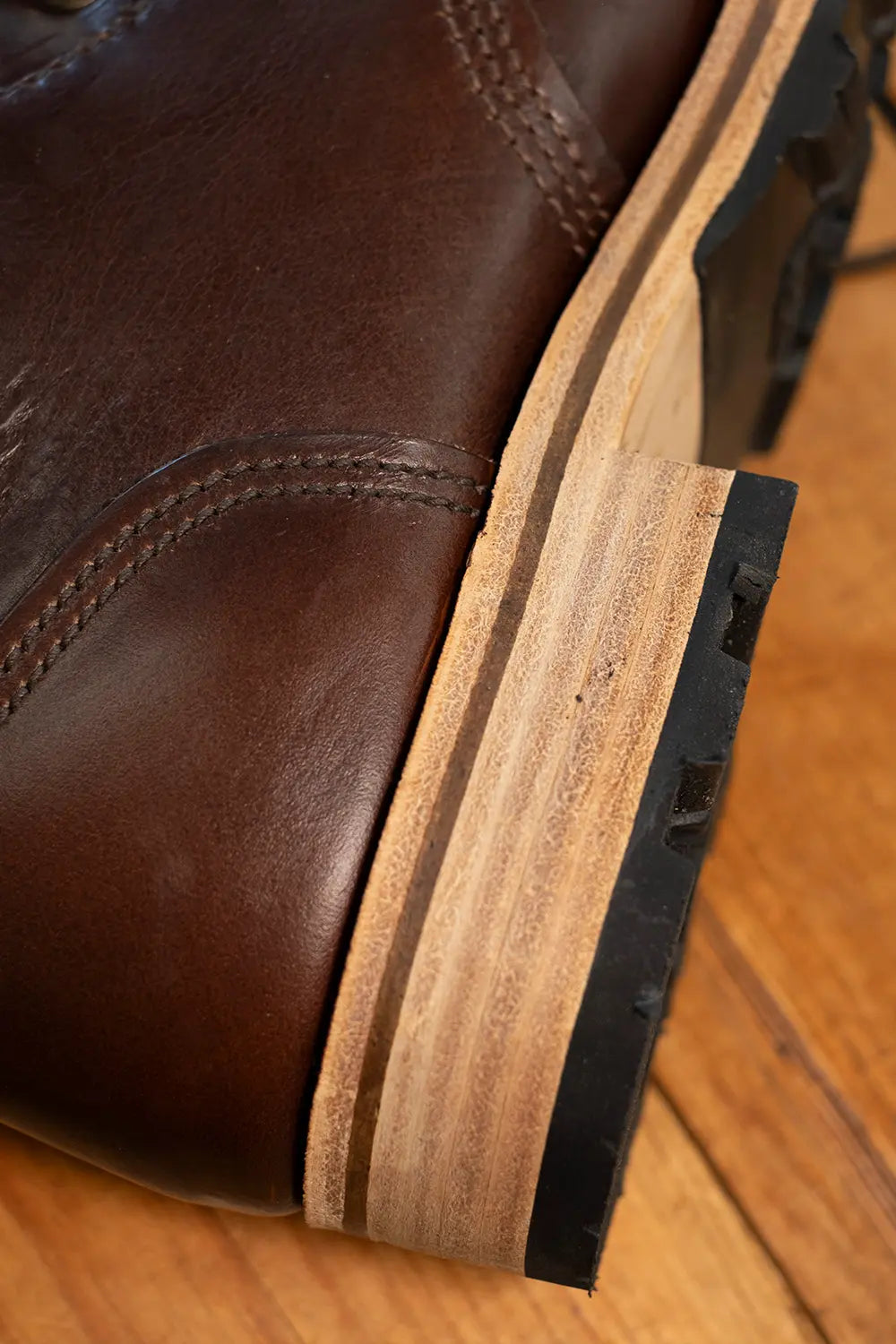 Close-up of a dark brown leather boot showing the layered sole, featuring a natural wood inlay and a black rubber outsole, resting on a wooden floor.