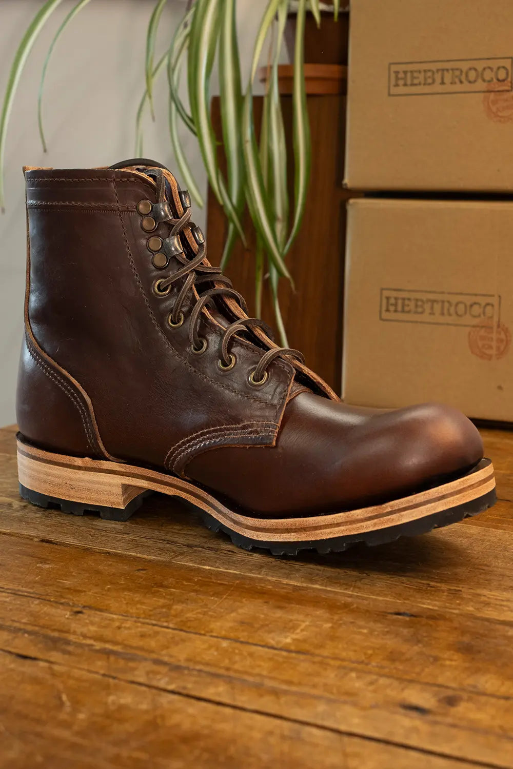 A dark brown leather lace-up work boot with a stacked wood-look heel, resting on a wooden surface with cardboard boxes and a plant in the background.