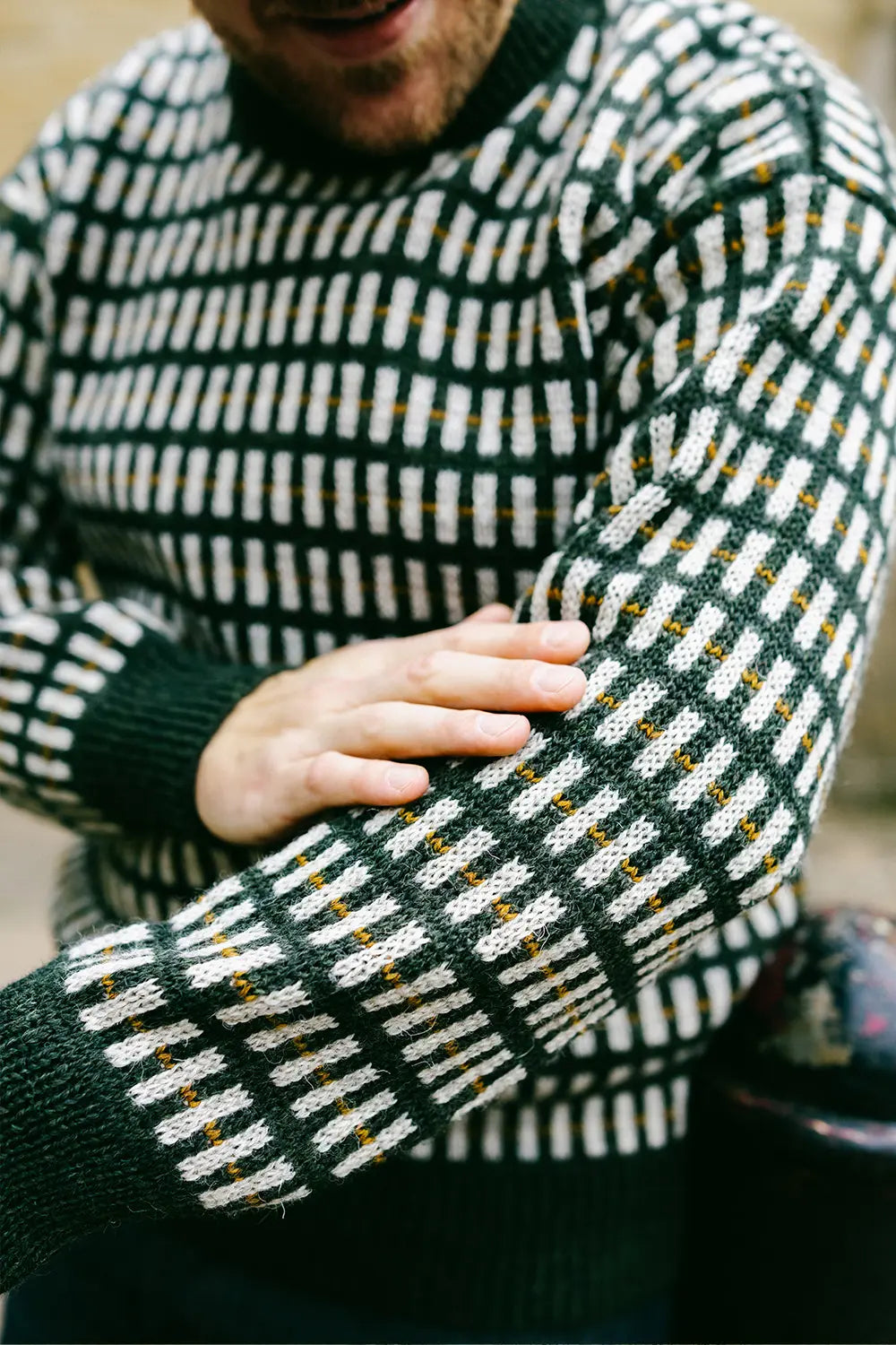 Close-up of a man wearing a dark green and white patterned knit jumper, showing the texture of the fabric and his hand resting on his arm.
