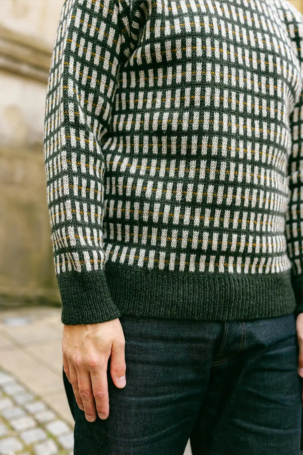 A close-up of a person wearing a dark green knit sweater with a cream and tan geometric check pattern, paired with dark blue jeans.