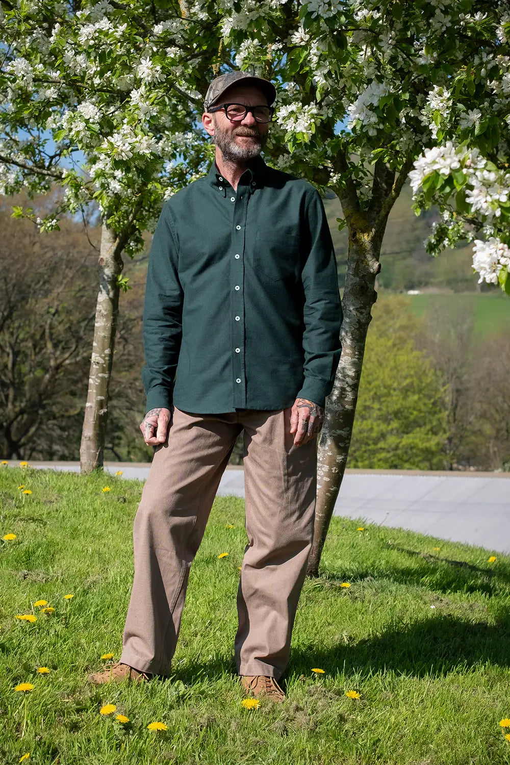 A man wearing a dark green long-sleeve button-up shirt, tan pants, and a dark cap stands outdoors on a grassy lawn under flowering trees.