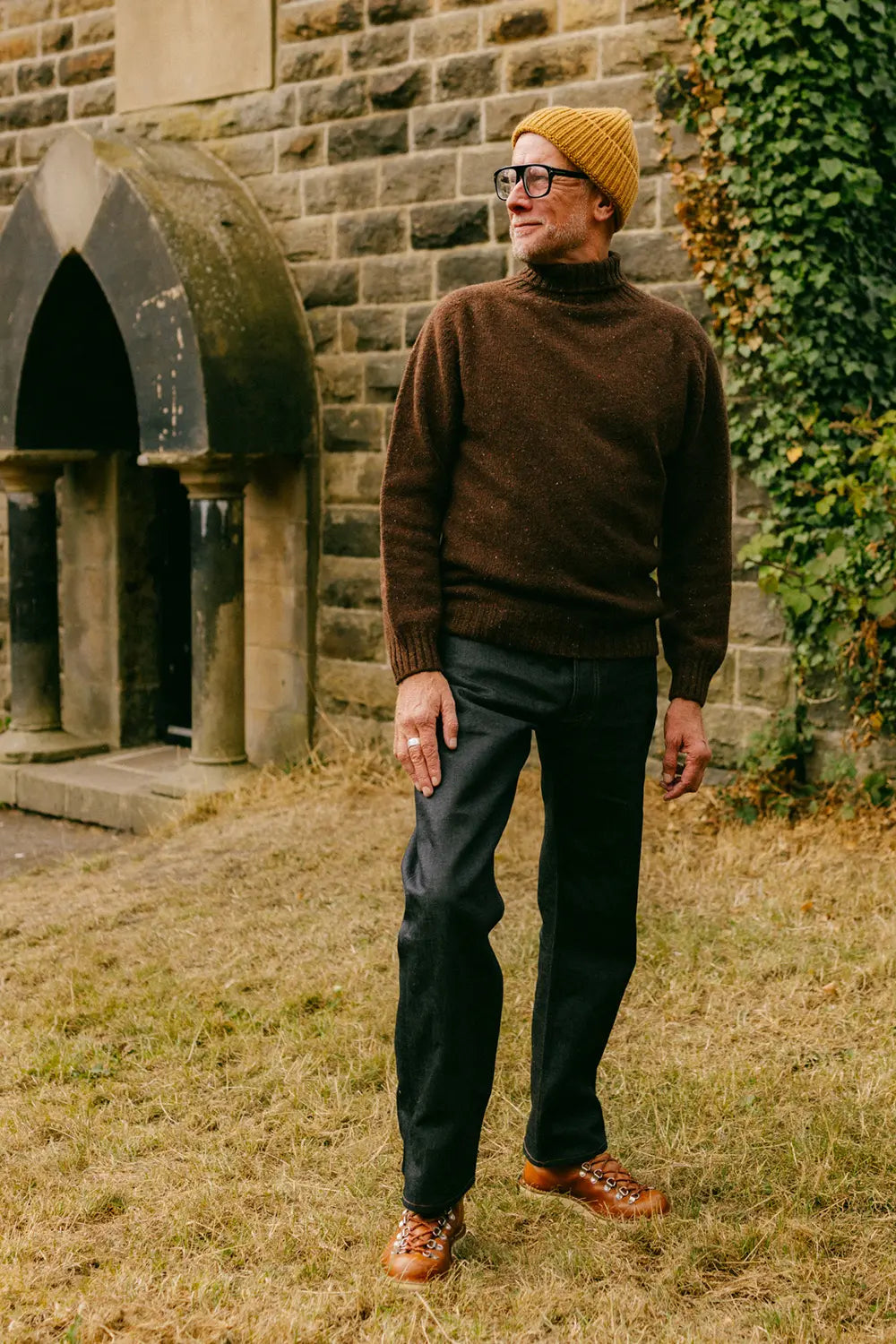 A man wearing a brown turtleneck sweater, dark trousers, a mustard yellow knit beanie, and brown shoes stands outdoors in front of a stone building with ivy.
