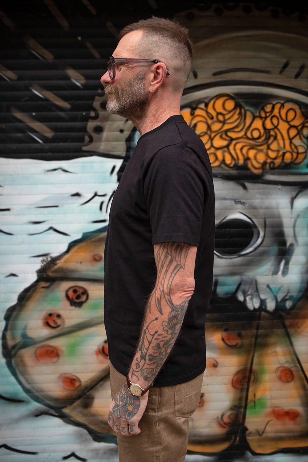 A man with glasses, a beard, and tattoos stands in profile wearing a black t-shirt and khaki pants, set against a colorful graffiti wall.