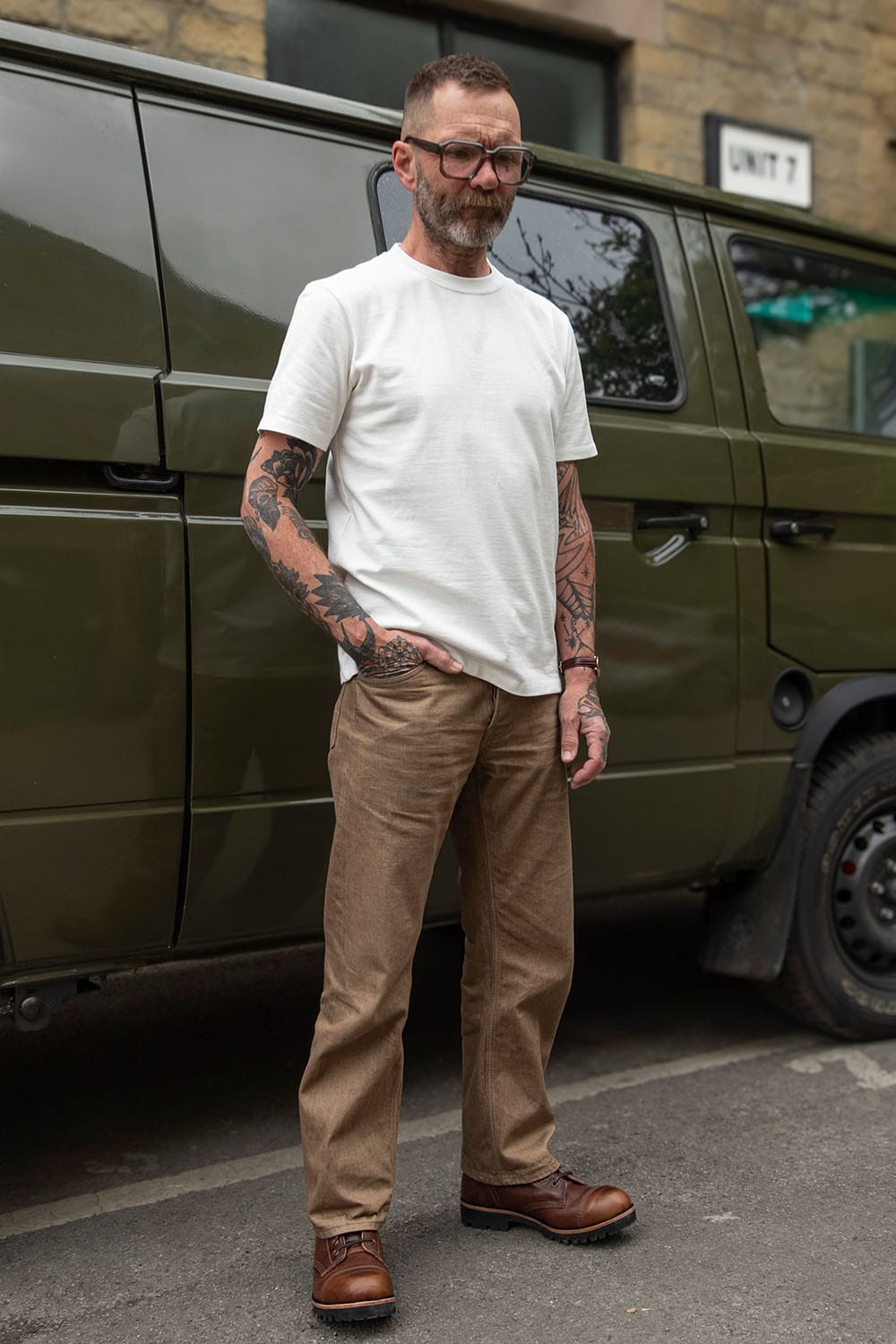 A man with tattoos and glasses stands in front of an olive green van, wearing a white crew-neck t-shirt, brown chinos, and brown leather shoes.
