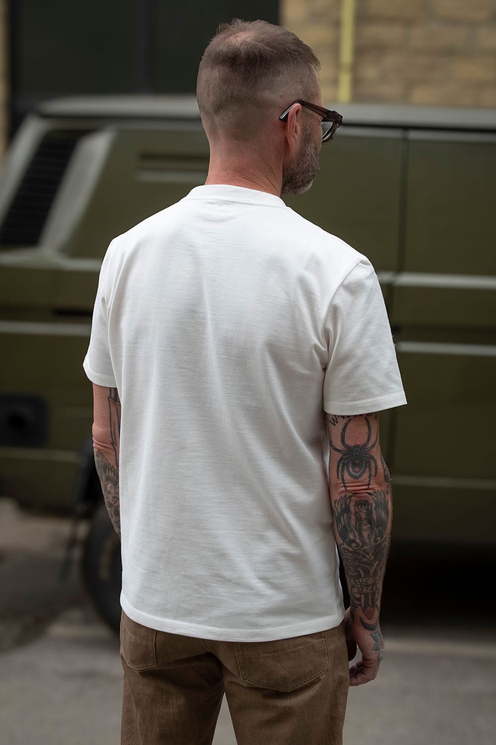 A man viewed from behind wearing a plain white short-sleeve crewneck t-shirt and brown trousers, standing outdoors in front of a dark green military-style vehicle.