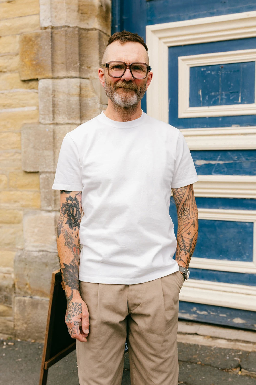 A man with tattoos and glasses wearing a plain white crew-neck t-shirt and beige trousers, standing outdoors in front of a blue and white wooden door and a stone wall.