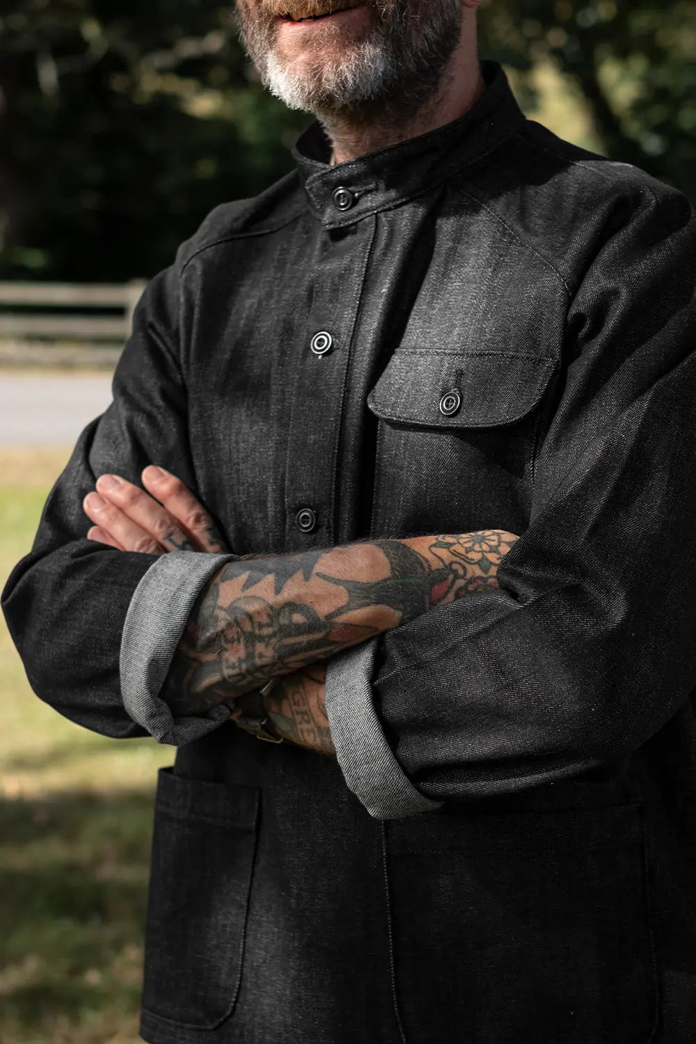 A close-up of a man wearing a dark charcoal gray textured button-up shirt jacket with a high collar and rolled-up sleeves showing a light gray lining, set against a blurred outdoor background.