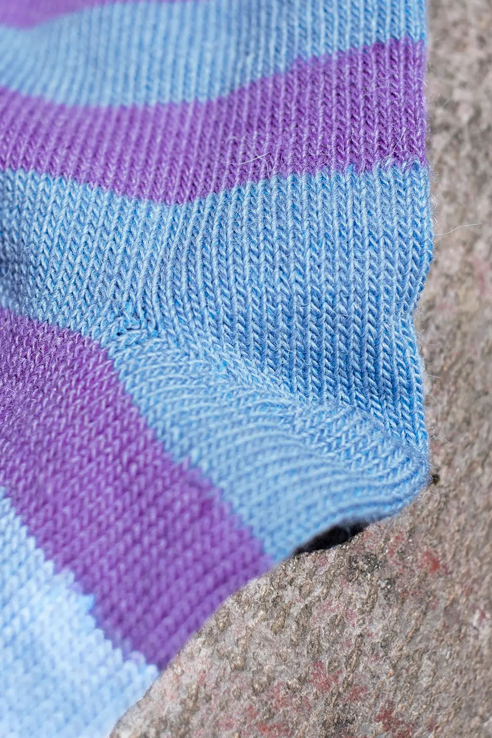 A close-up of a knitted sock with horizontal stripes in light blue and purple, resting against a textured gray stone surface.