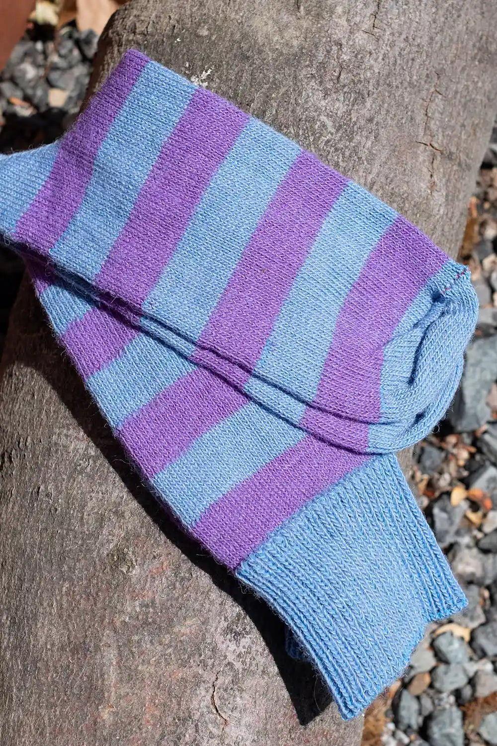 A close-up of a folded knit sock with alternating light blue and purple stripes, resting on a textured brown log outdoors with gravel visible in the background.