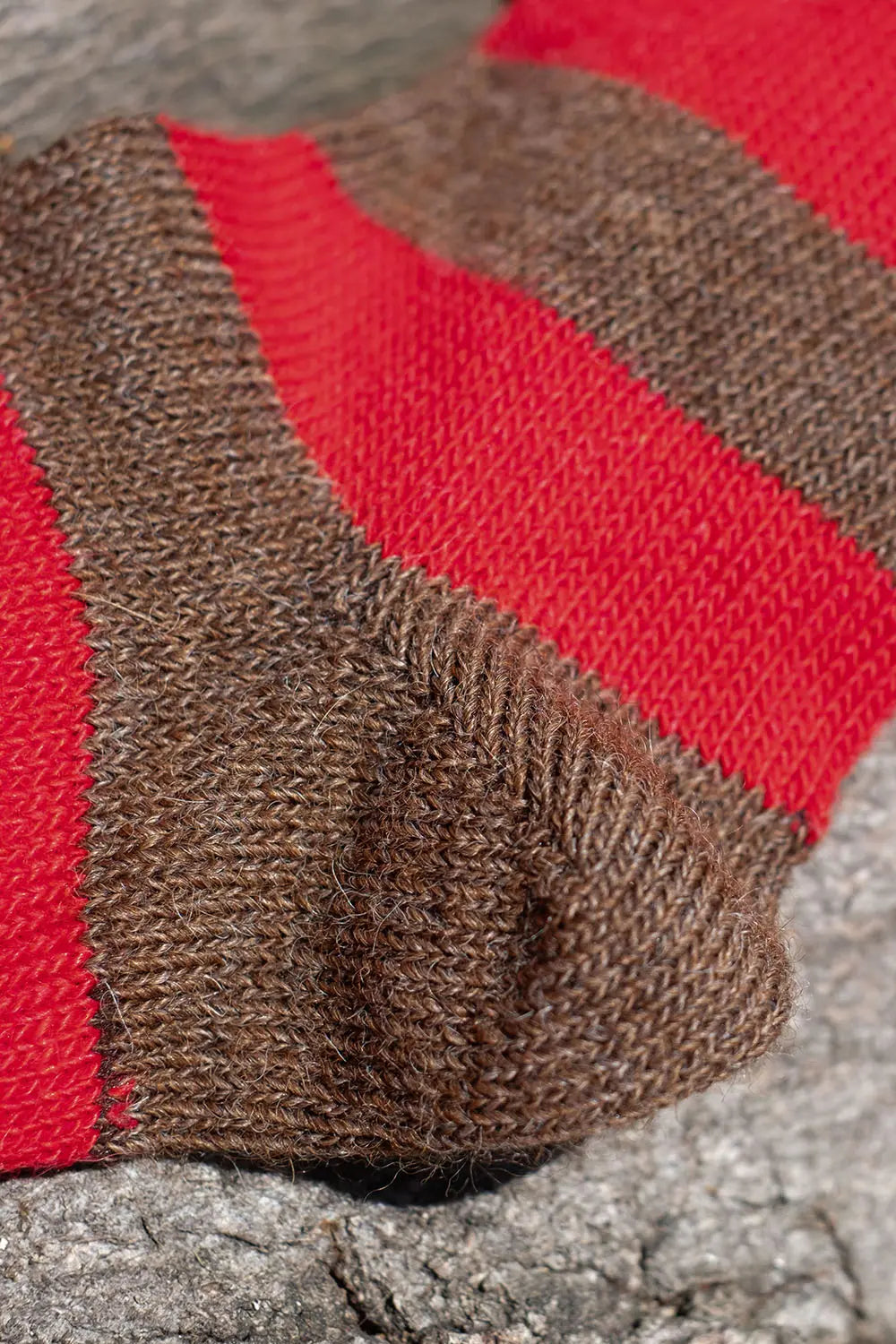 A close-up of a hand-knit sock with alternating thick stripes of brown and red wool, resting on a textured gray stone surface.