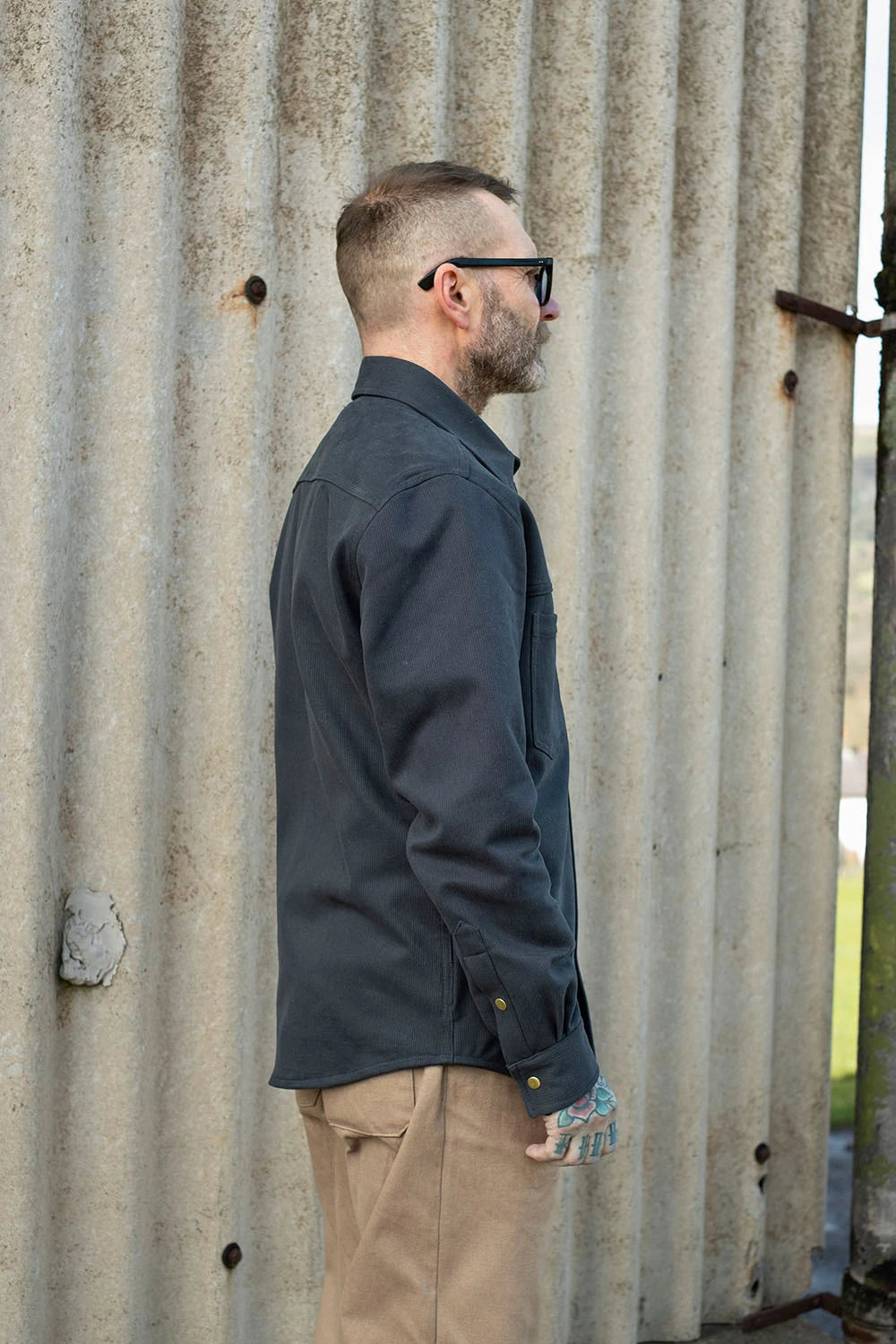 A man in profile wearing a navy bedford cord ironshirt and tan pants, standing in front of a corrugated metal wall.