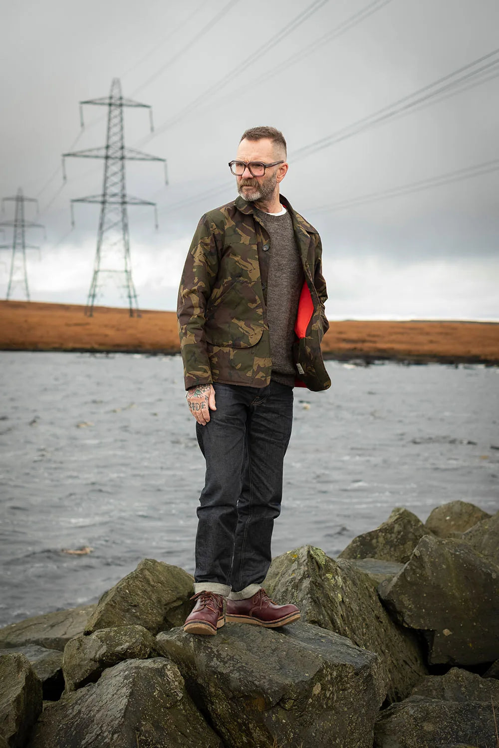 Ed wearing a green camouflage Jacksnipe jacket, dark gray sweatshirt, dark jeans, and brown shoes stands on large rocks by the water with power lines in the background.