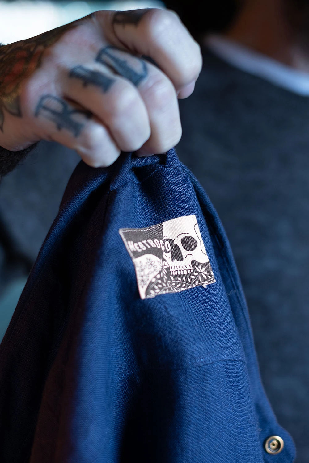 A close-up of a person's tattooed hand holding up a navy blue shirt jacket to show a sewn-on patch label that reads 'HEBTROCO'.