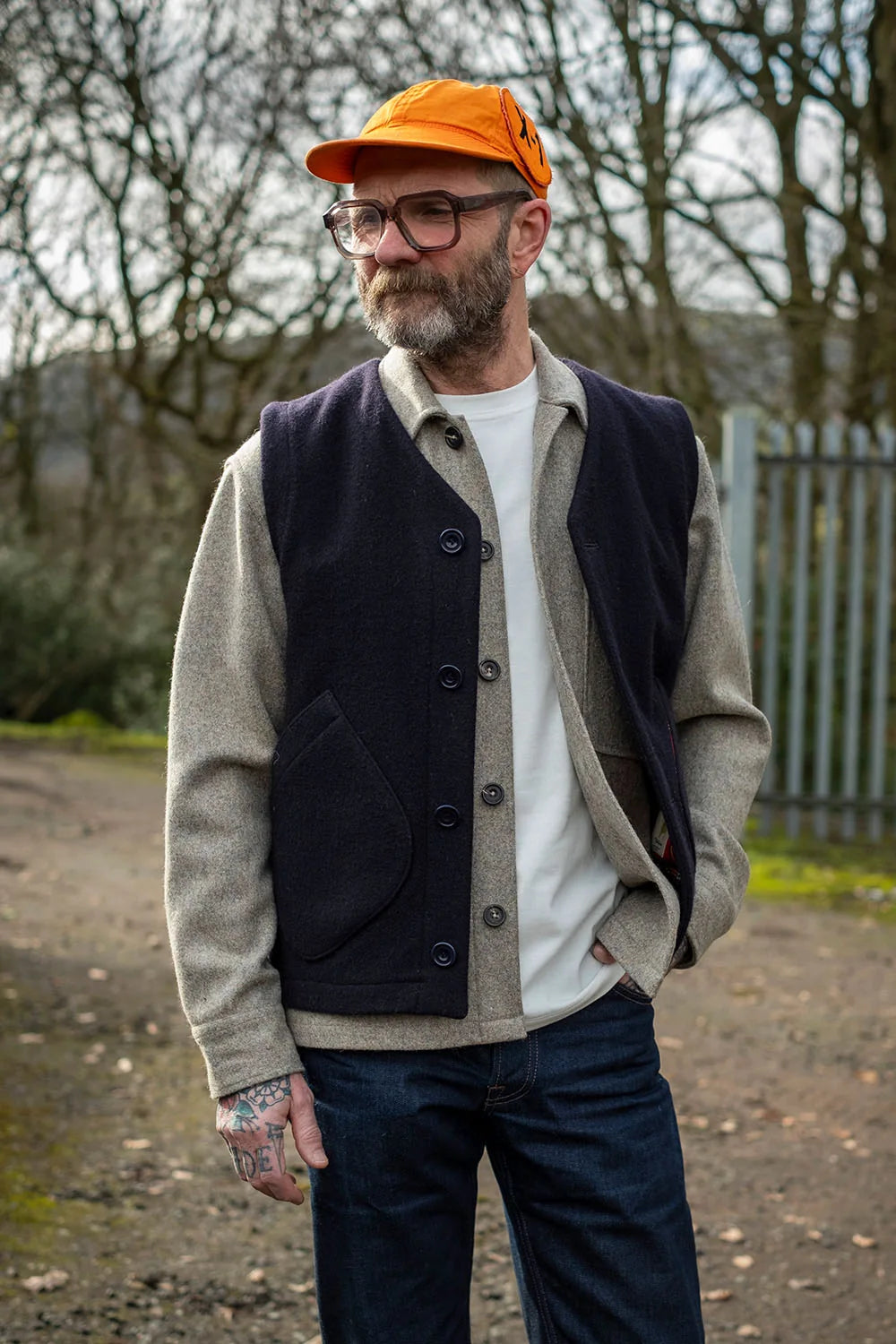 A man wearing a dark navy wool button-front vest over a beige knit cardigan, a white t-shirt, dark blue jeans, and an orange cap, standing outdoors on a dirt path.