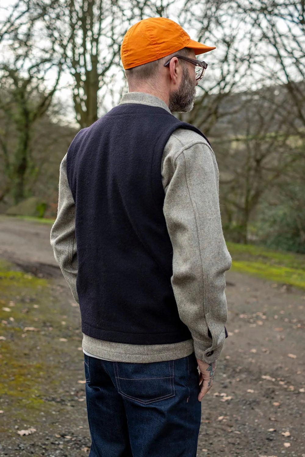A man wearing a dark navy knit sweater vest over a light gray long-sleeve shirt, blue jeans, and an orange baseball cap, standing outdoors on a dirt path with trees in the background.