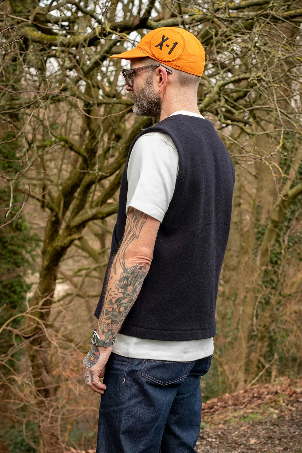 A man wearing a dark navy sleeveless knit sweater vest over a white t-shirt, blue jeans, and an orange cap with 'X1' printed on it, standing outdoors in a wooded area.