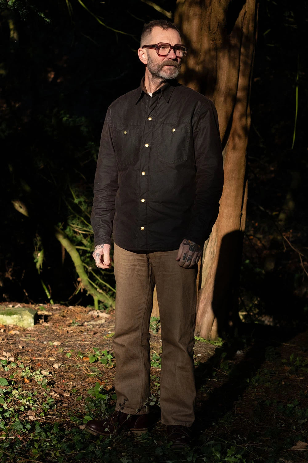 A man wearing a black long-sleeve button-up shirt and brown pants standing outdoors in a dark, wooded environment.