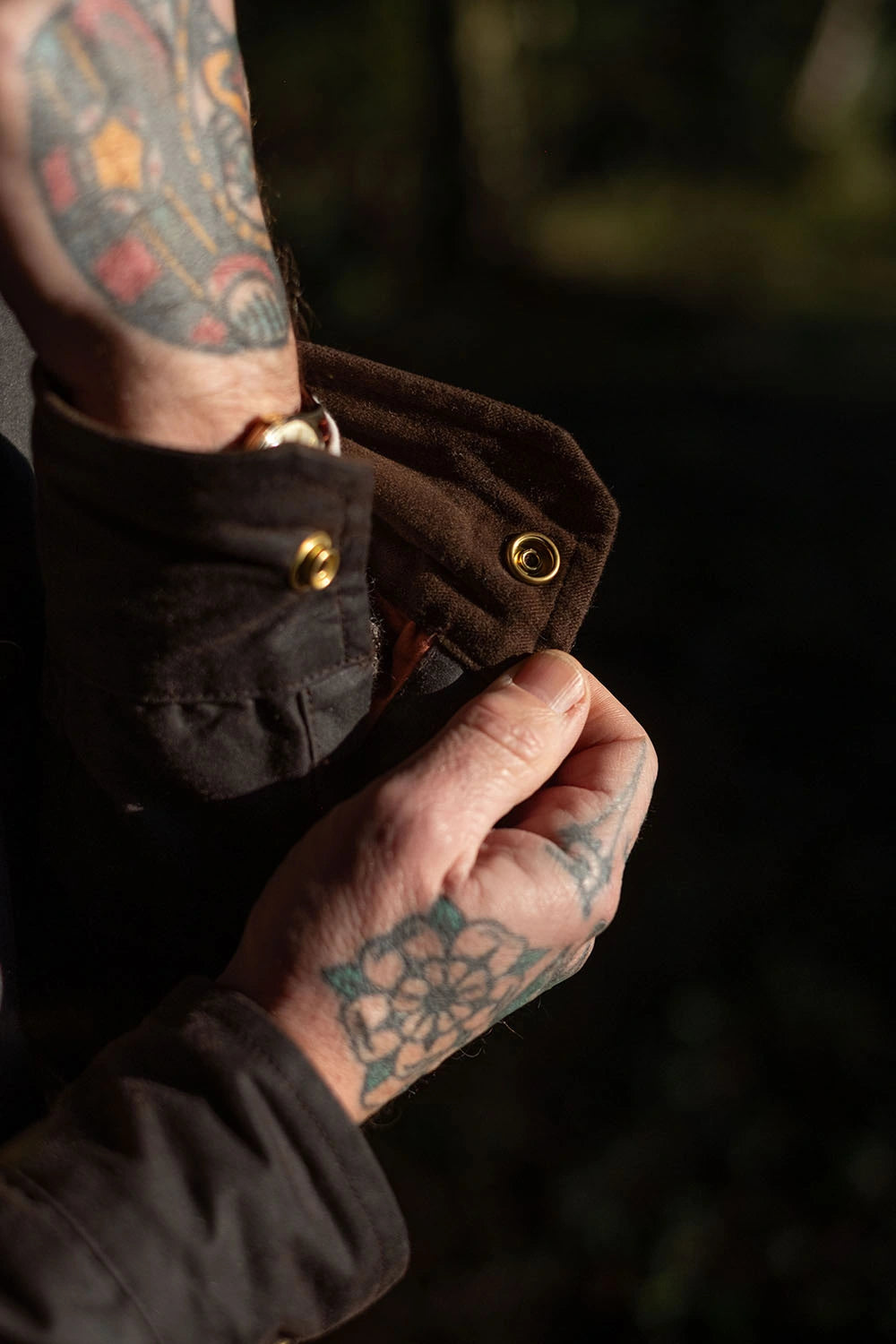 Close-up of a person's tattooed hands and forearm adjusting a dark brown jacket cuff with brass snap buttons, set against a dark, blurred outdoor background.