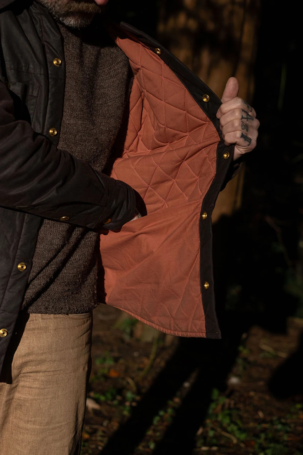 A close-up of a person wearing a black quilted jacket and opening the front to show a rust-orange quilted lining. The jacket has gold-tone snap buttons and is styled over a brown knit sweater and beige trousers in a dark, outdoor setting.