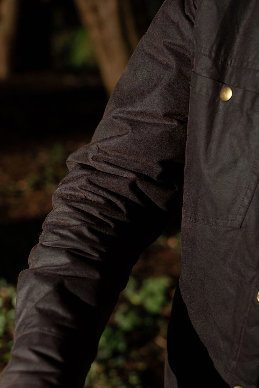 Close-up of a person wearing a dark charcoal gray waxed cotton jacket outdoors, showing the textured sleeve and a brass button on the chest pocket.