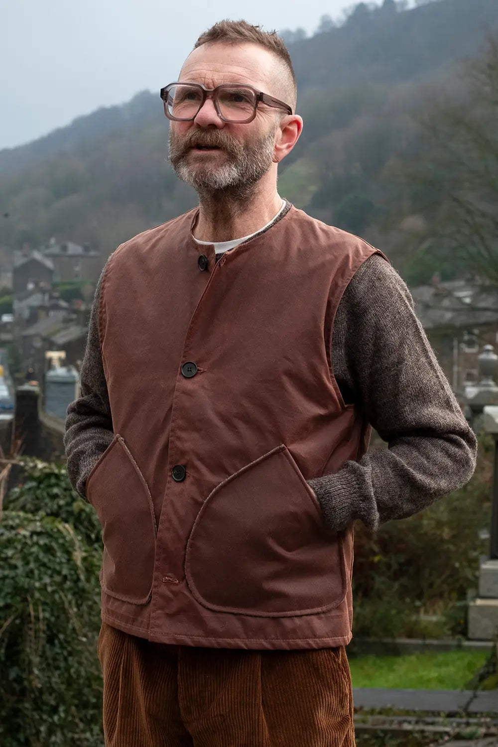 Ed wearing a rascal vest over a gray knit jumper,  standing outdoors with a blurred background of hills and buildings.