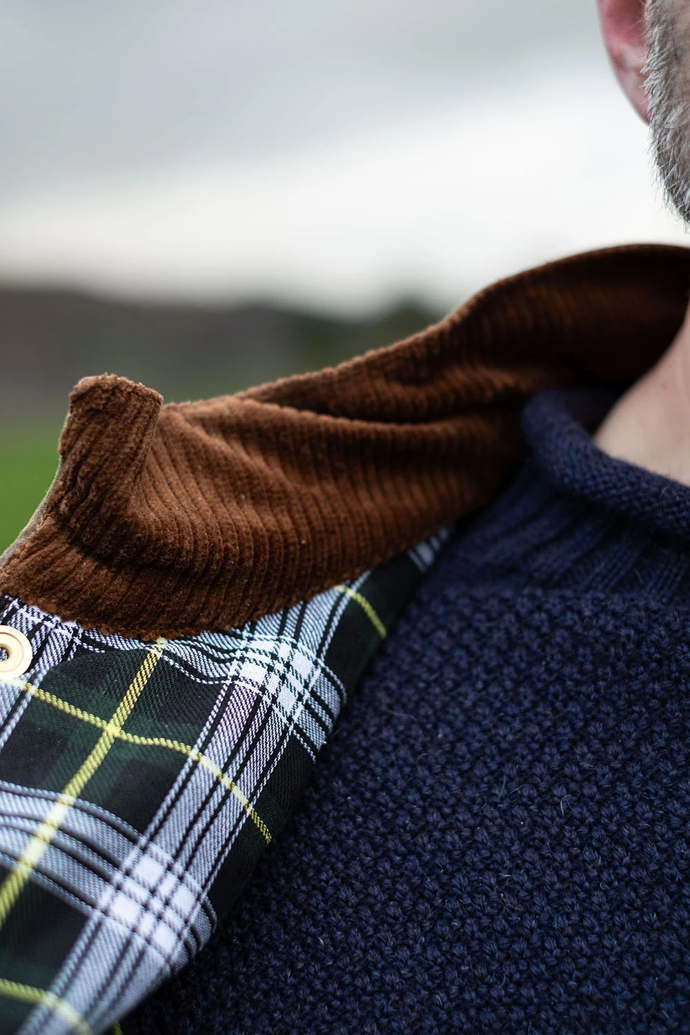 Close-up of a person wearing a navy knit sweater and a brown corduroy collar layered over a black, white, and green plaid shirt.
