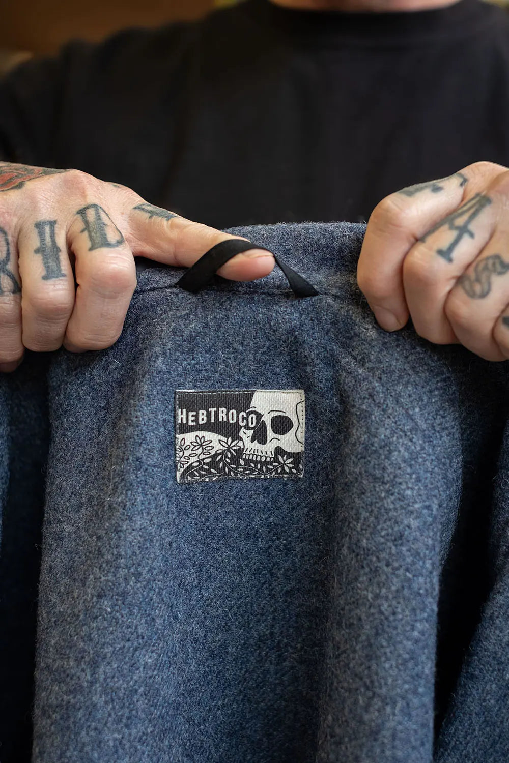 A close-up of a person holding up a dark blue wool jacket, showing the brand patch 'HEBTROCO' on the inside. The person's tattooed hands are visible on either side of the jacket.