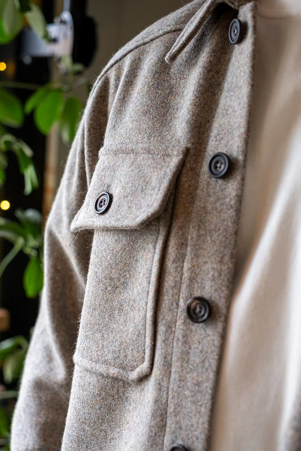 Close-up of a USB37 wool overshirt with dark buttons, worn over a cream shirt, with blurred green foliage in the background.